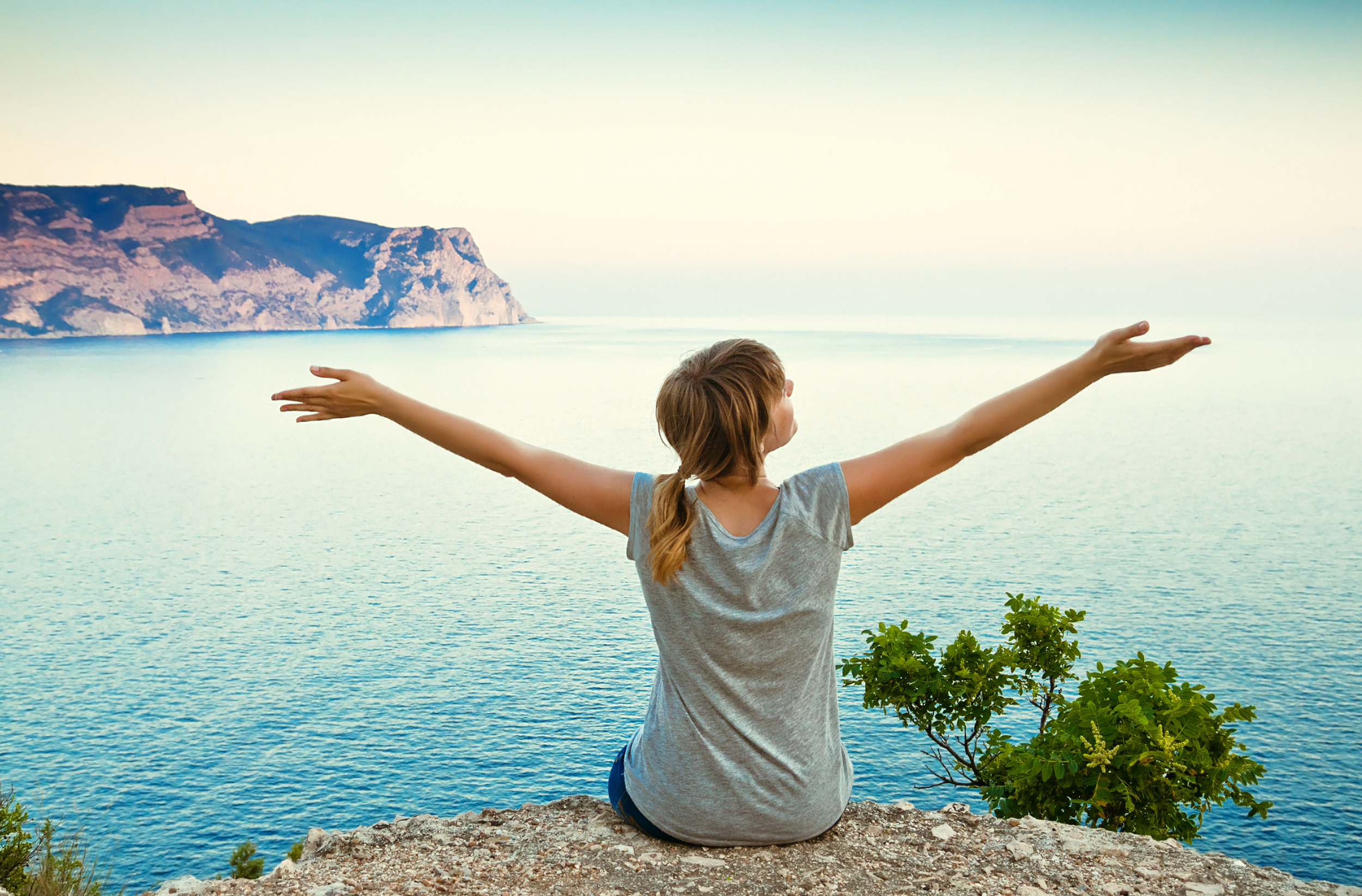 girl sits on a hill with raised hands and looking at the sea